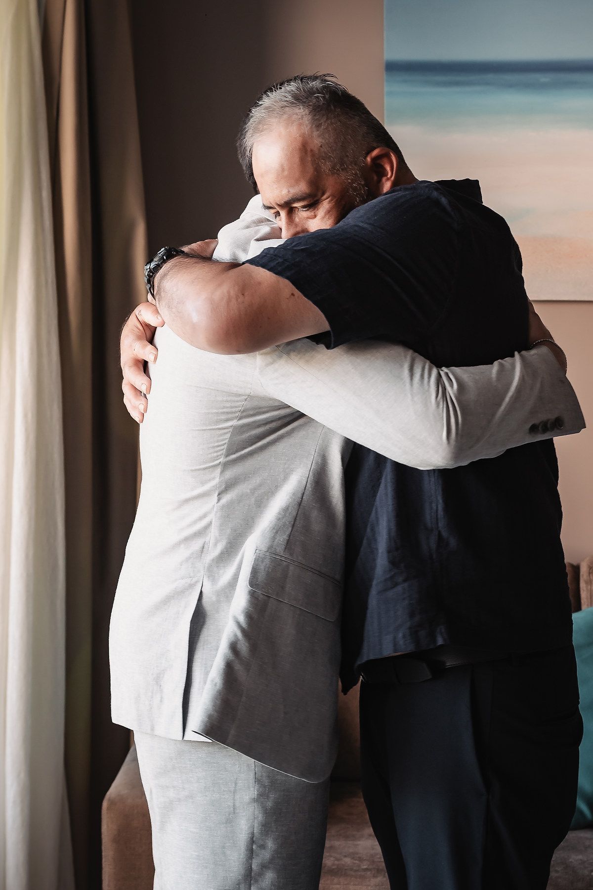Two middle-aged men sharing an emotional embrace in a warmly lit room with ocean artwork in the background.