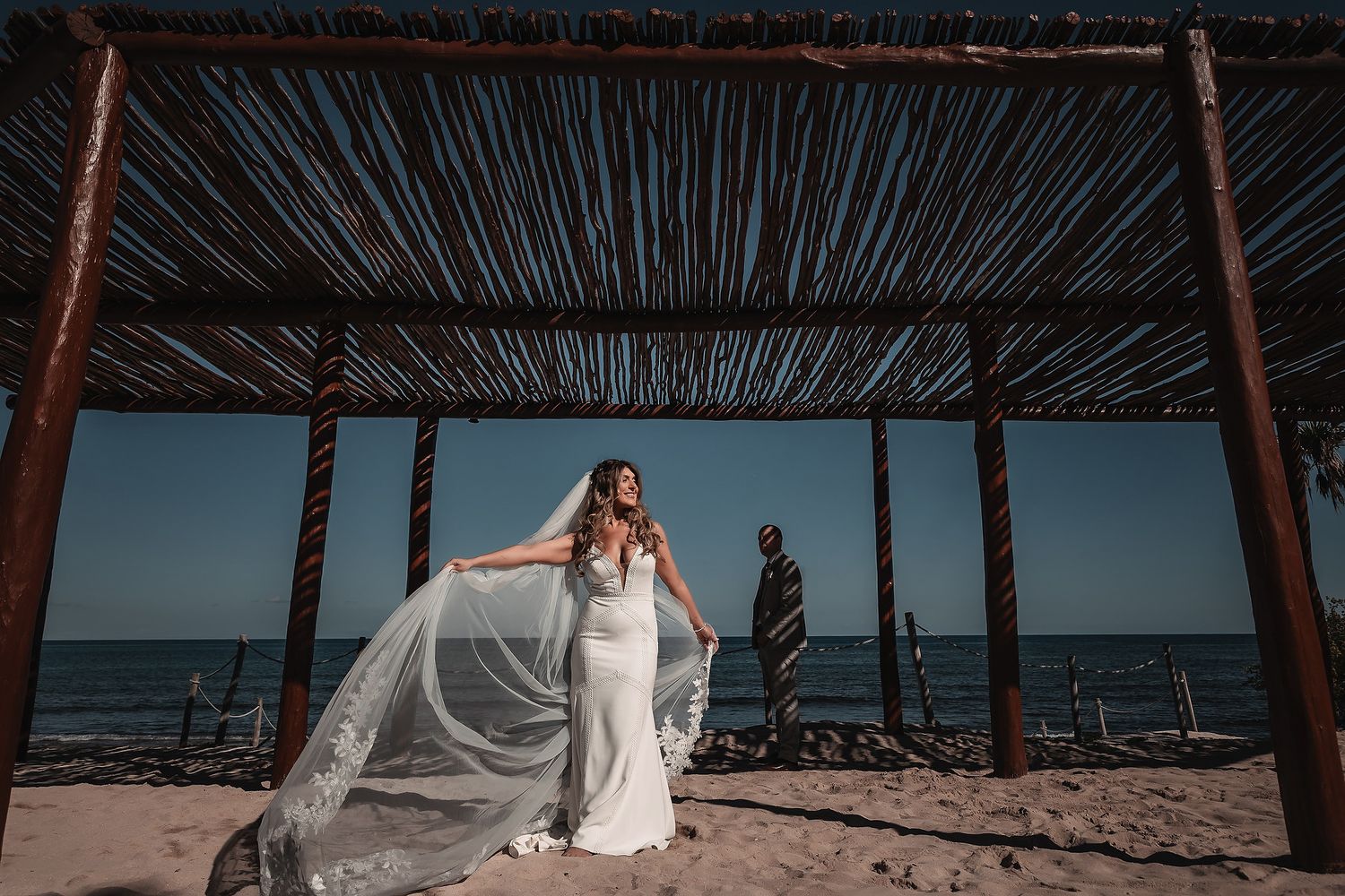 Bride in white gown with flowing veil on sandy beach under rustic wooden pergola with ocean backdrop.