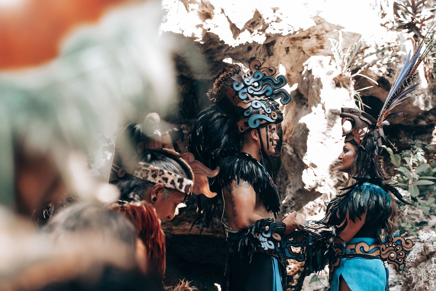 People dressed in traditional Mayan ceremonial attire with feathered headdresses gather near ancient stone ruins.