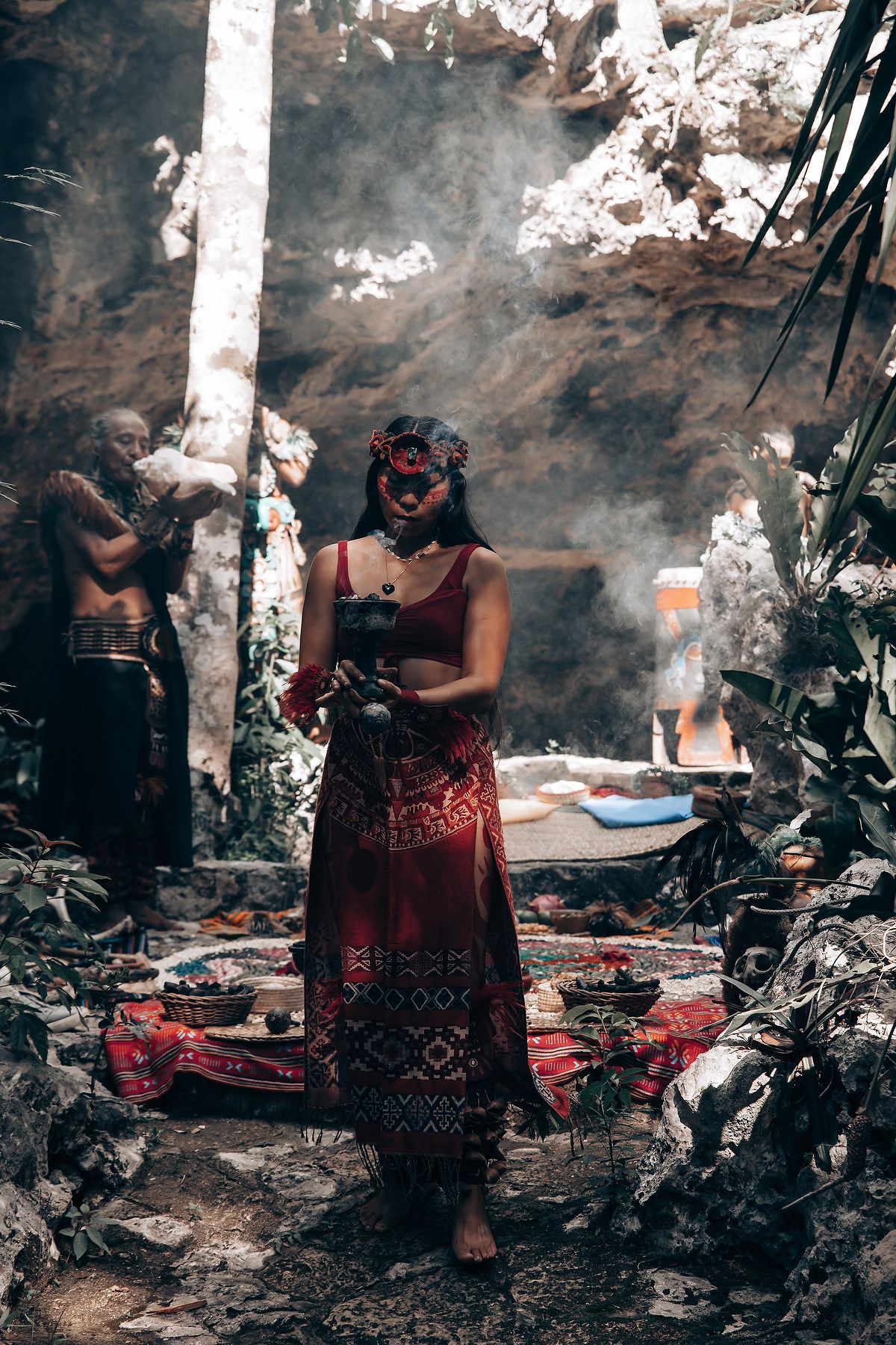 Woman in red traditional Mayan dress holding ritual object in smoky ceremonial cave setting.