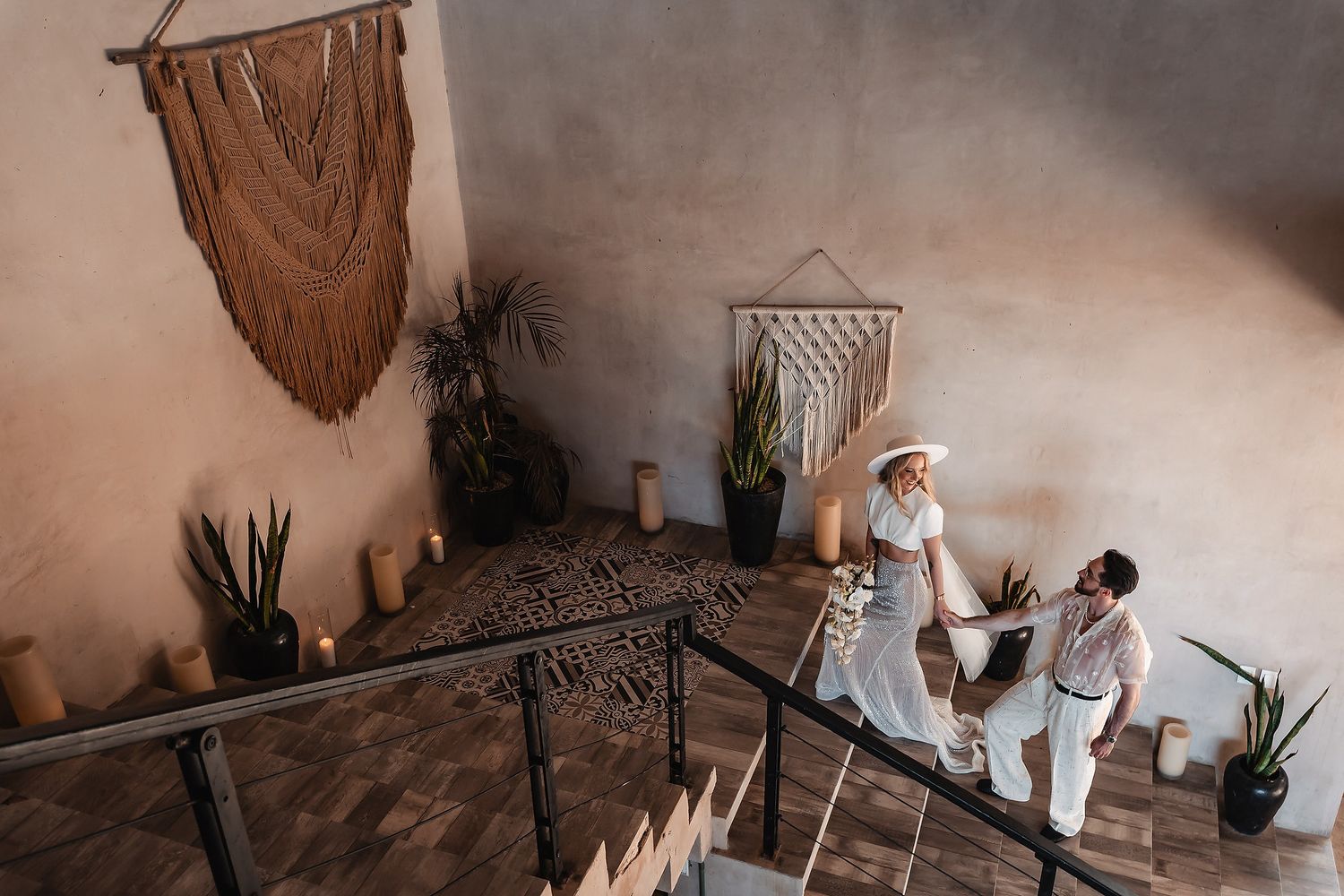 Couple in boho wedding attire walking up stairs with macrame wall art and tropical plants in a rustic venue.