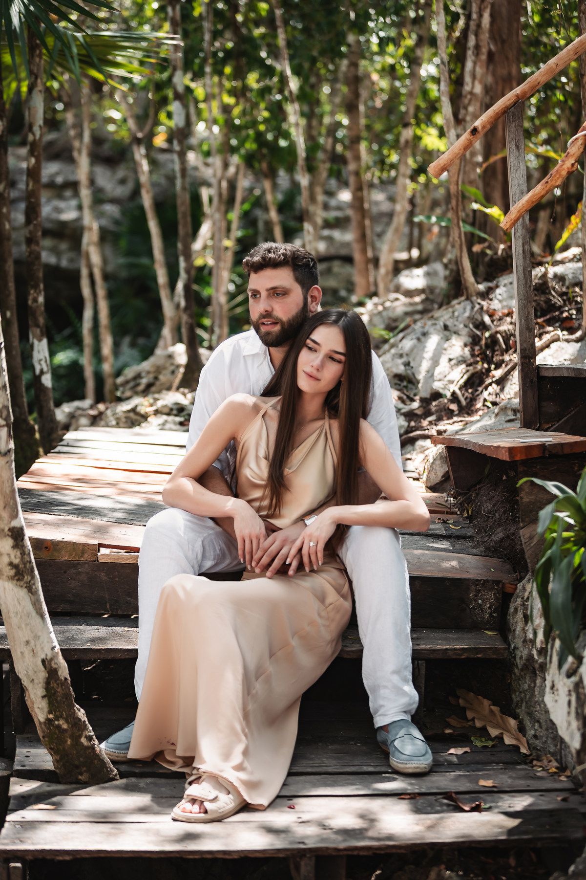 A couple sits together on wooden steps in a lush tropical forest, the woman in a beige dress leaning on the man.