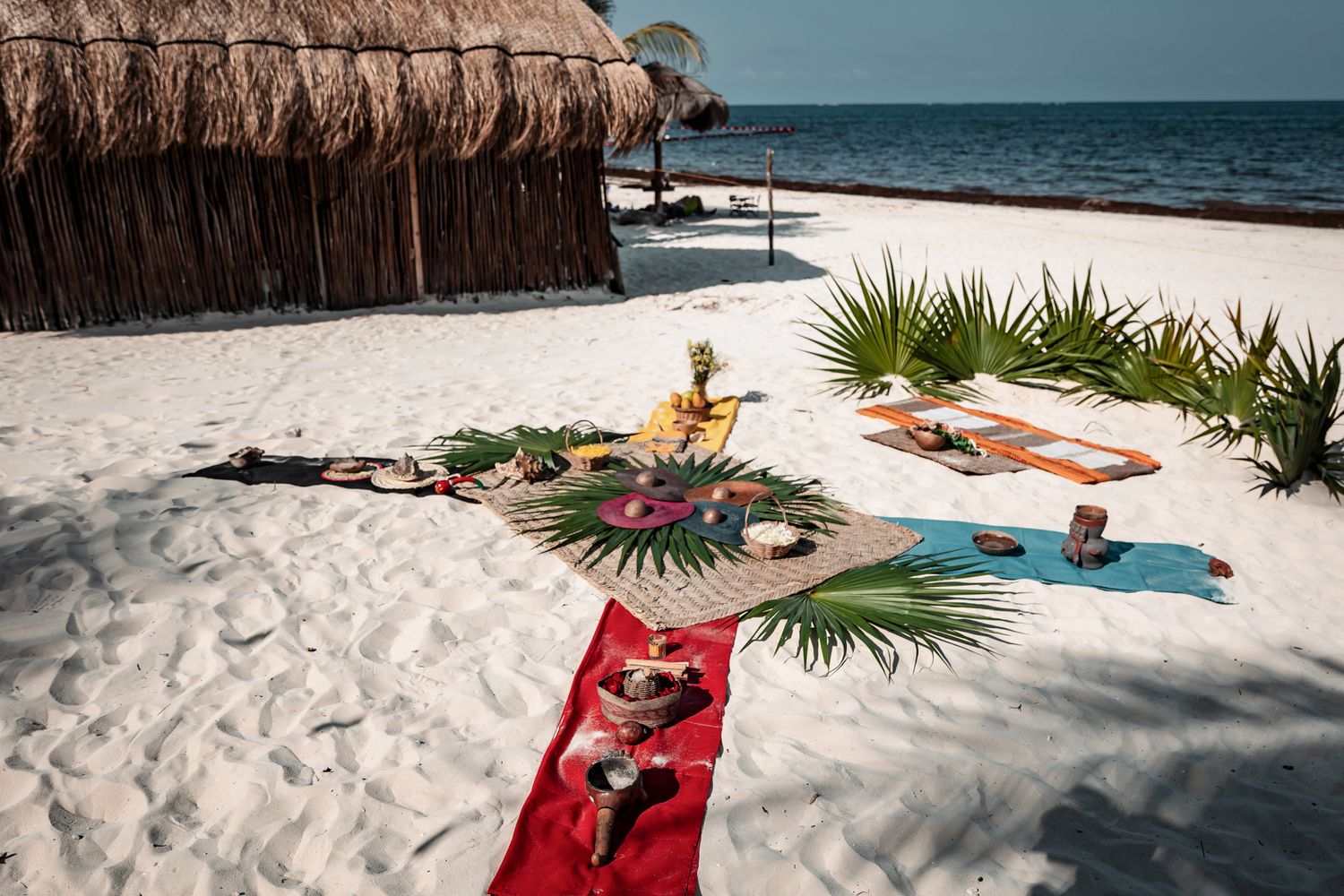 People practicing yoga on colorful mats on a white sandy beach near a thatched hut with tropical decor.