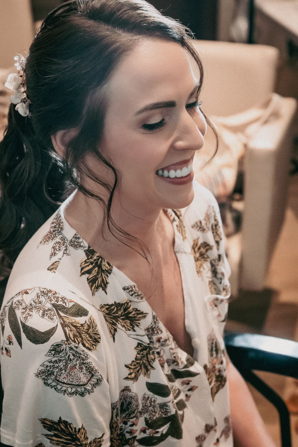 Smiling bride with dark hair adorned with floral accessories, wearing a white floral robe during wedding preparations.