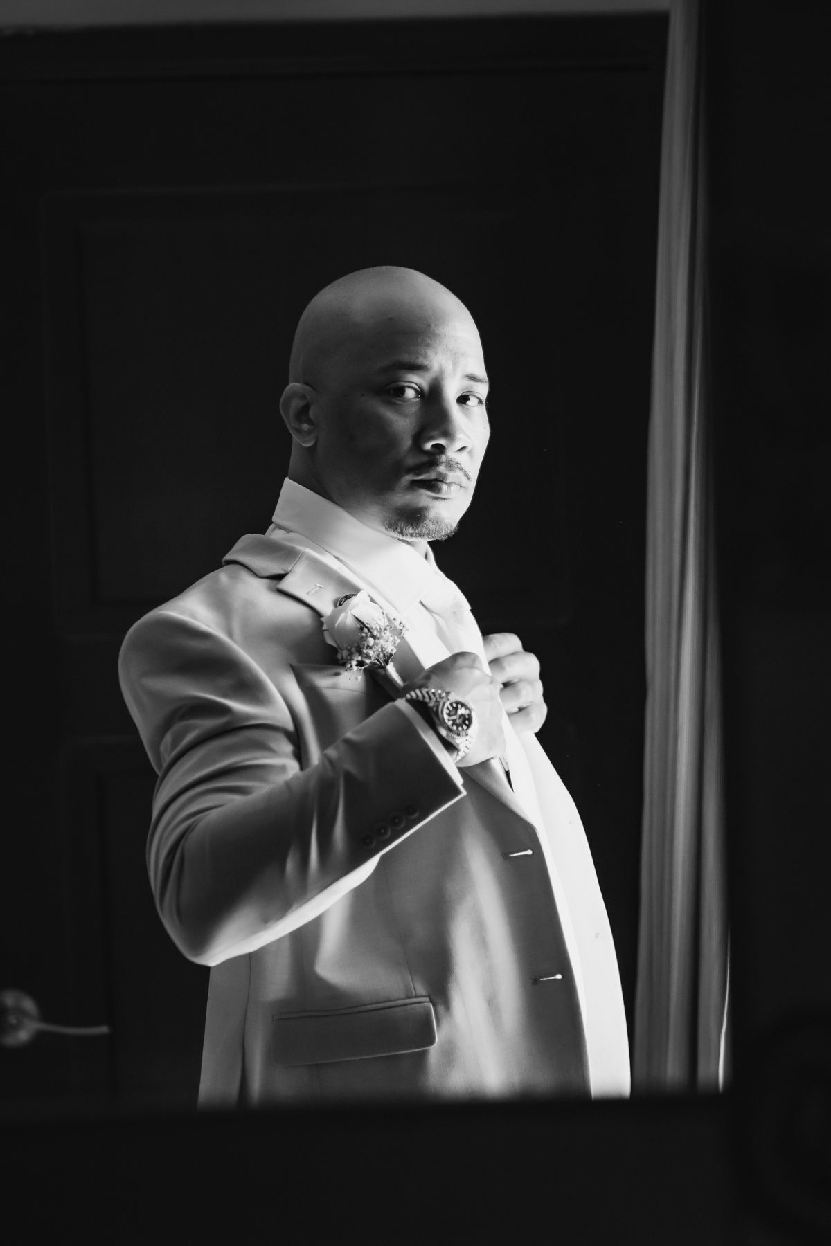 Bald groom adjusting boutonniere in dramatic black and white portrait near doorway.