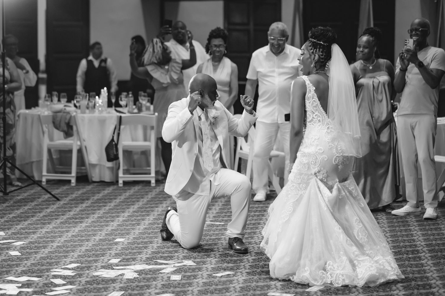 Groom kneels before bride during emotional first dance at wedding reception in black and white photo.