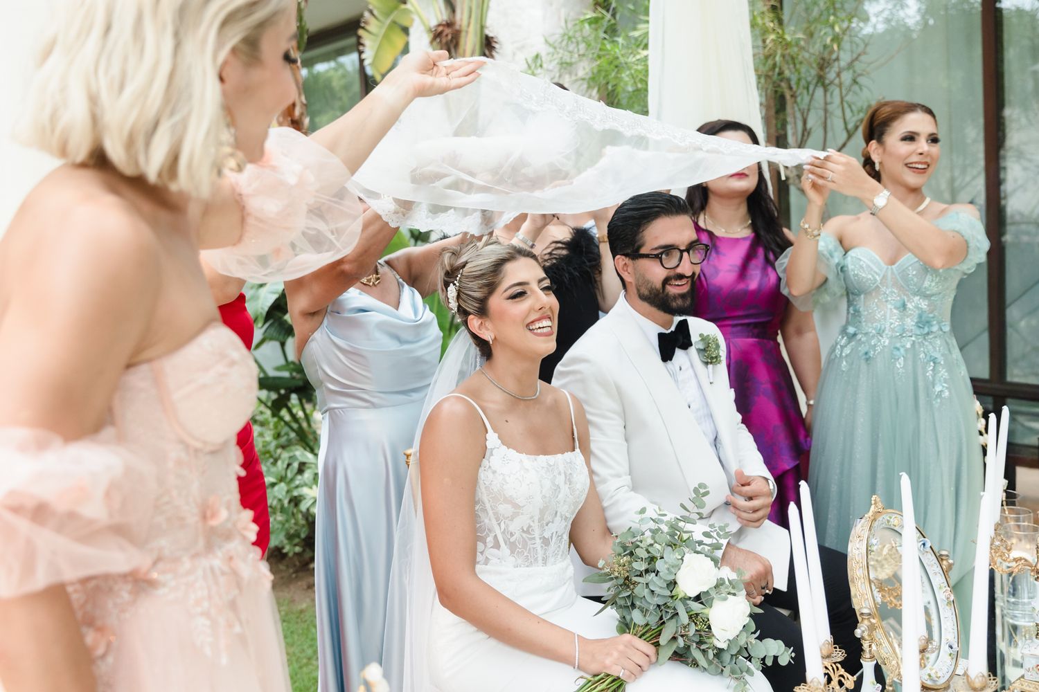 Bride and groom smile as bridesmaids in pastel gowns wave a veil overhead during a joyful outdoor wedding ceremony.