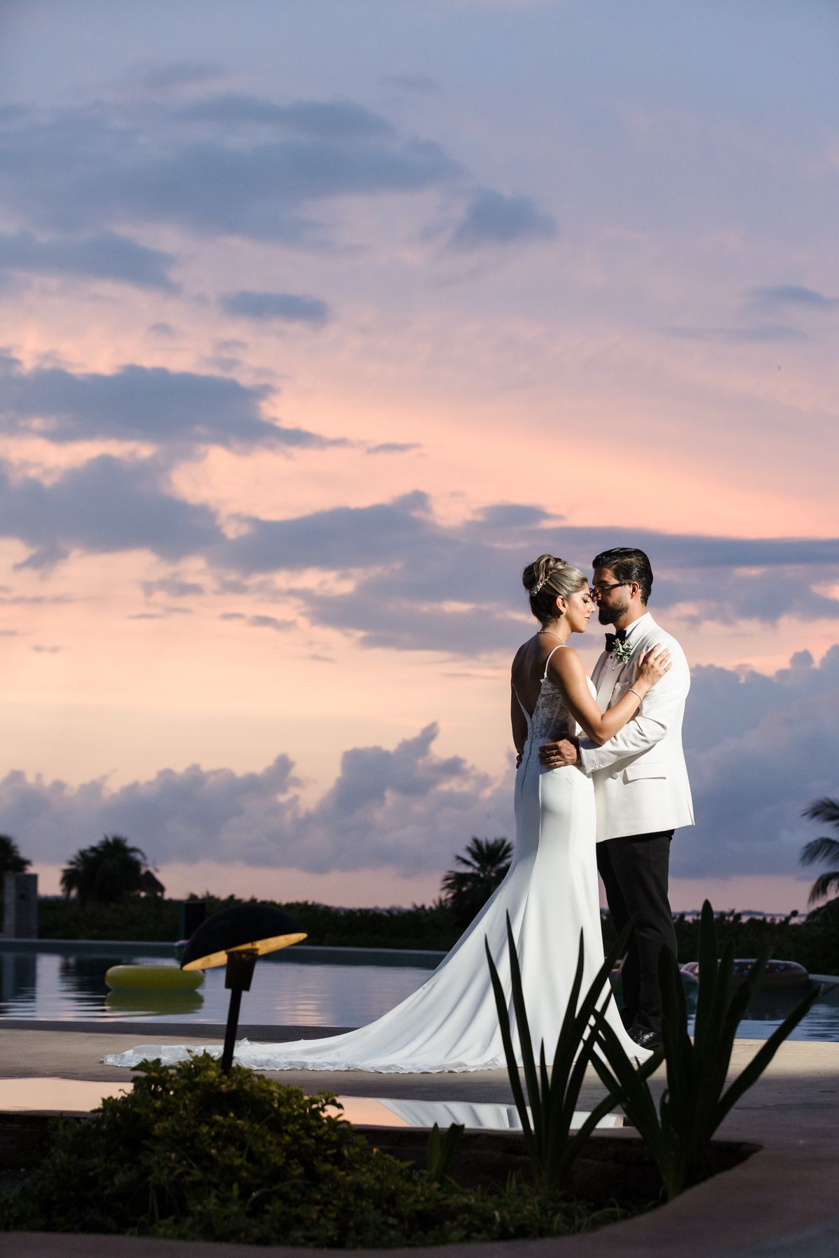 Bride and groom embrace poolside at sunset, pink and purple clouds reflecting over tropical resort waters.