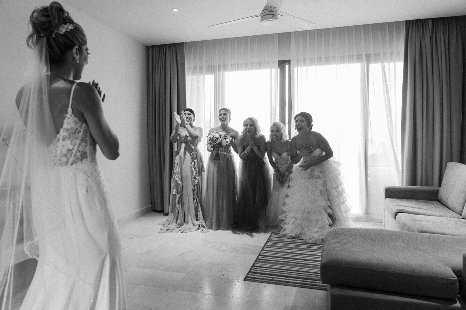 Bride reveals wedding dress to emotional bridesmaids in modern hotel room, captured in black and white photography.
