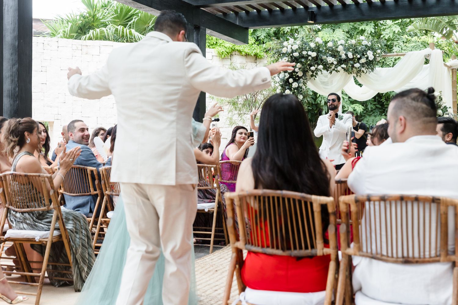 Groom in white suit dancing with guests seated at outdoor wedding reception under pergola.