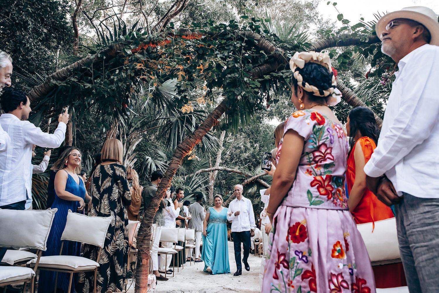 Outdoor wedding ceremony setup with tropical greenery arch and guests gathering under palm trees.