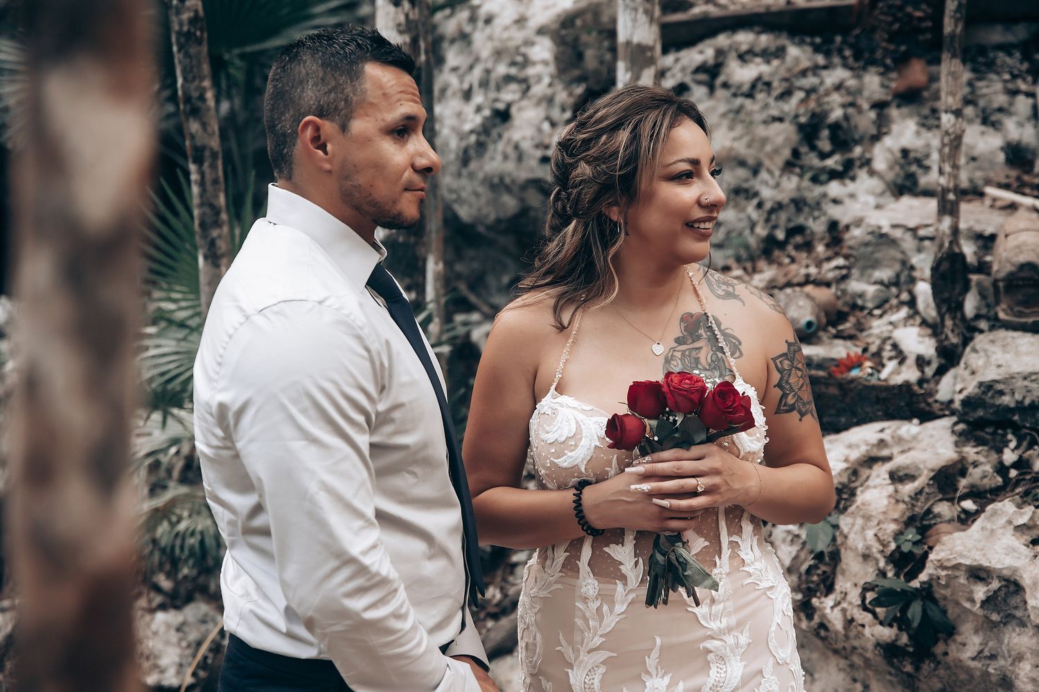 A bride in a lace gown holds a red bouquet while standing next to a groom in white against a stone wall backdrop.