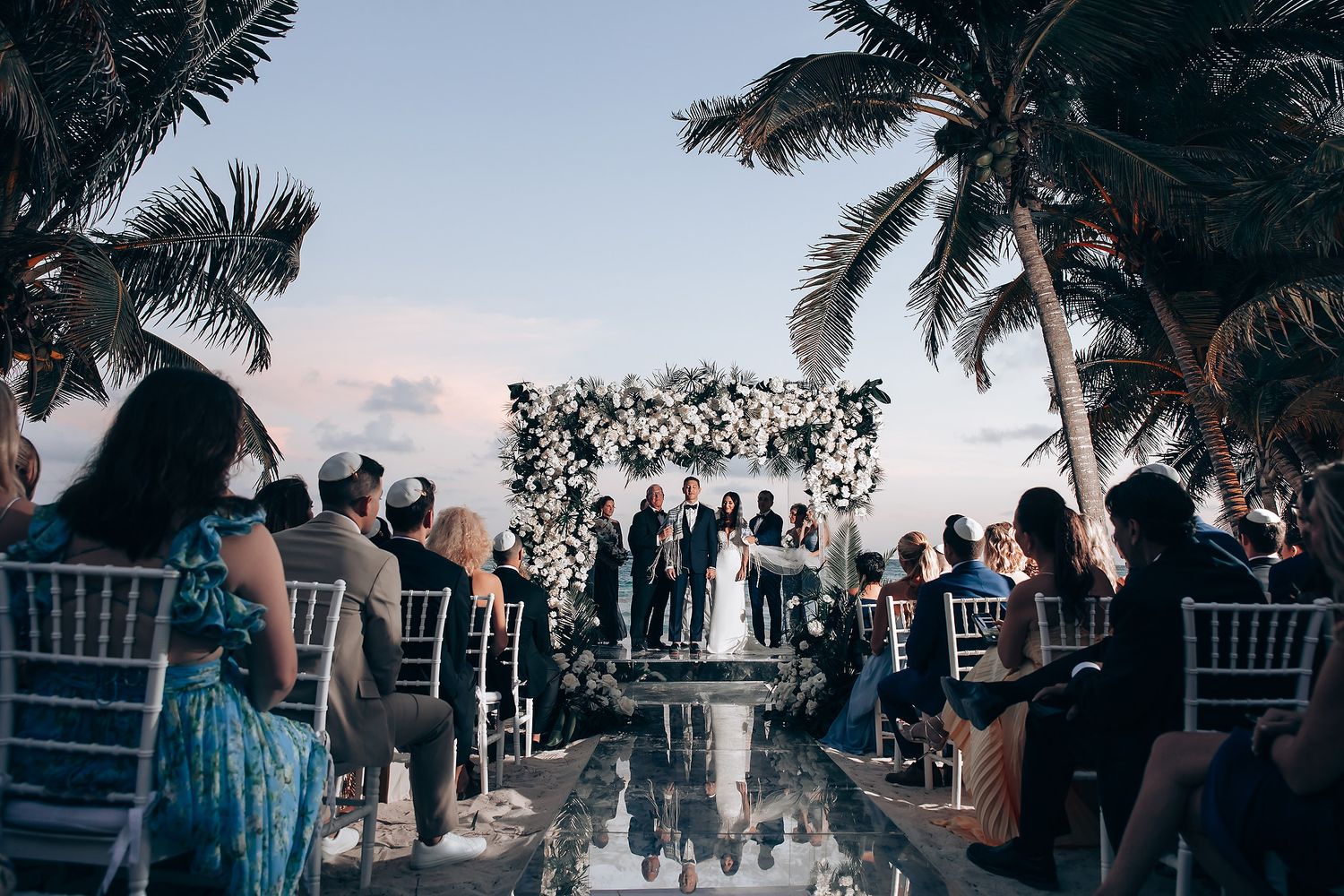 Outdoor wedding ceremony with guests seated among palm trees at sunset.