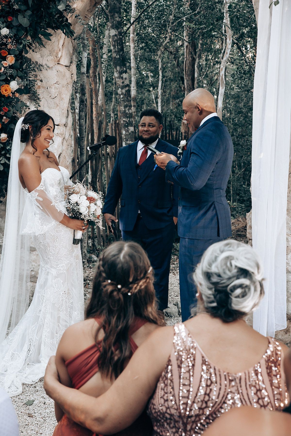 Wedding ceremony in progress under white tent with guests watching in a forest setting.