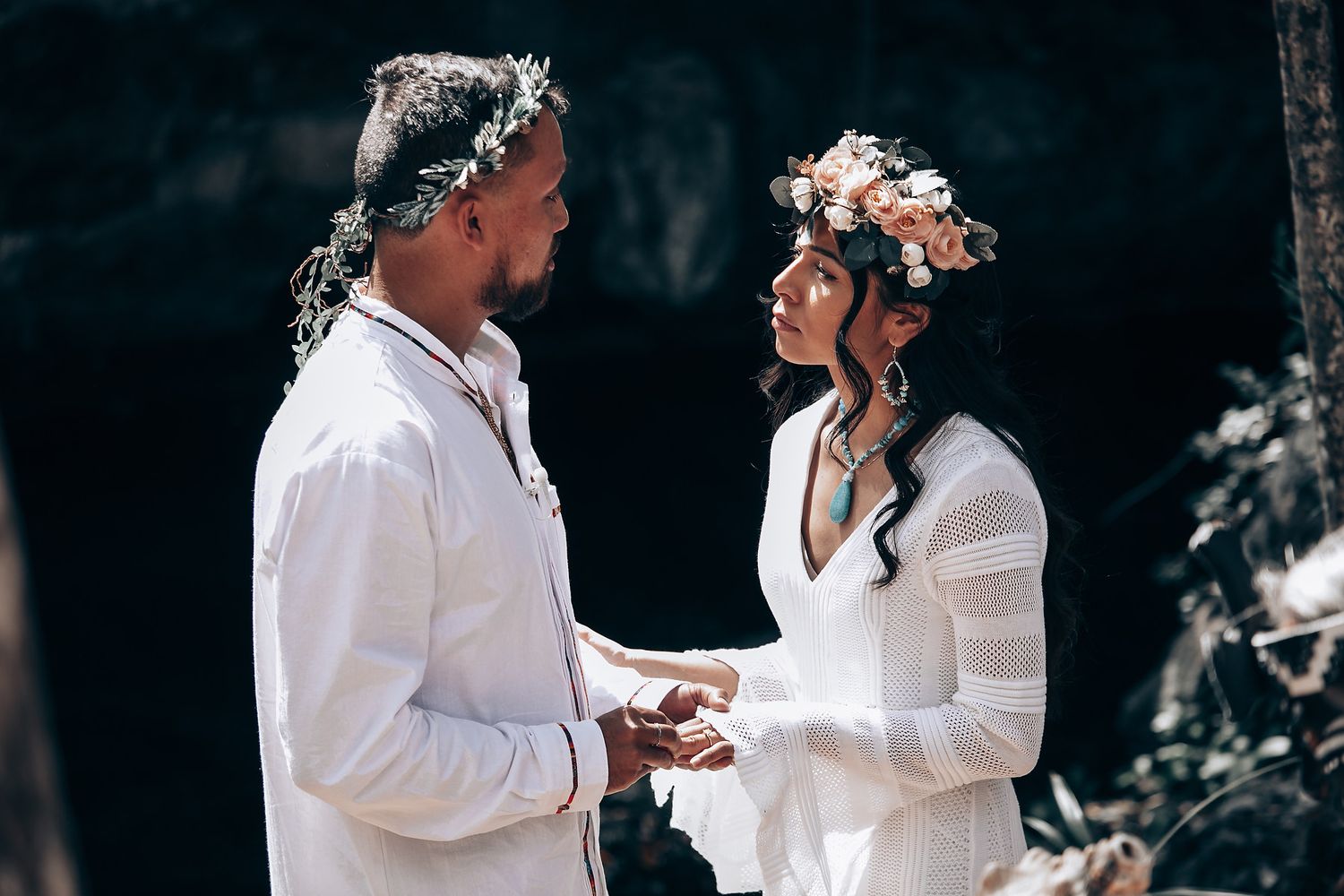 Tender moment between couple dressed in white against forest backdrop.