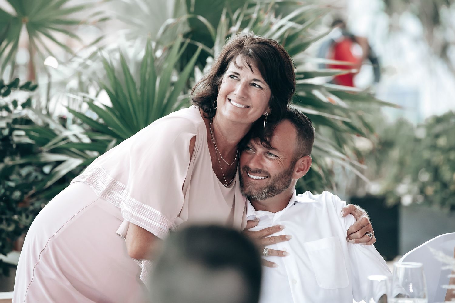 A couple embraces tenderly in an outdoor wedding setting with palm trees in the background.