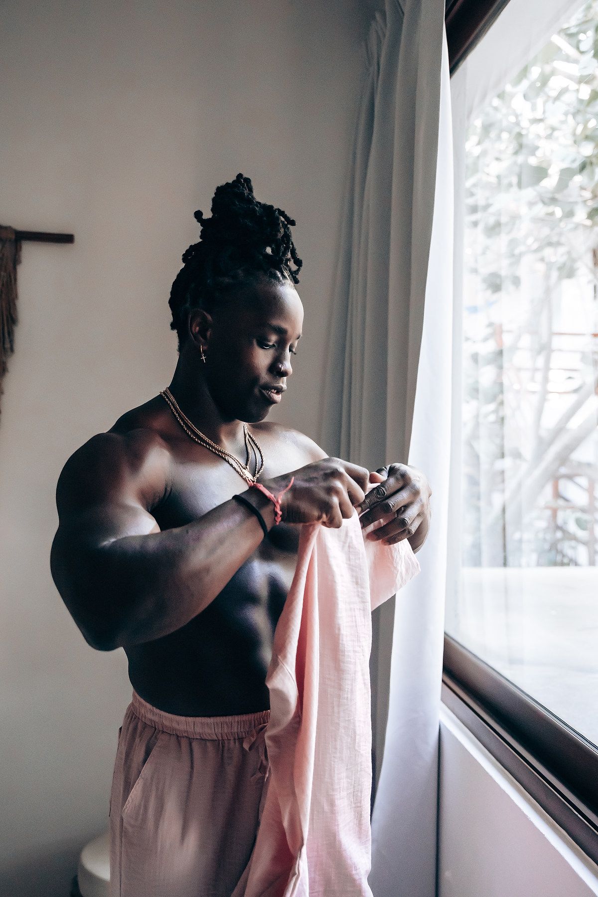 A shirtless man with dreadlocks stands in front of a window, holding a pink cloth.