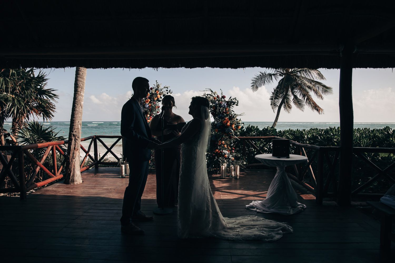 Silhouette of bride, groom, and officiant exchanging vows on tropical beachfront venue with ocean view.