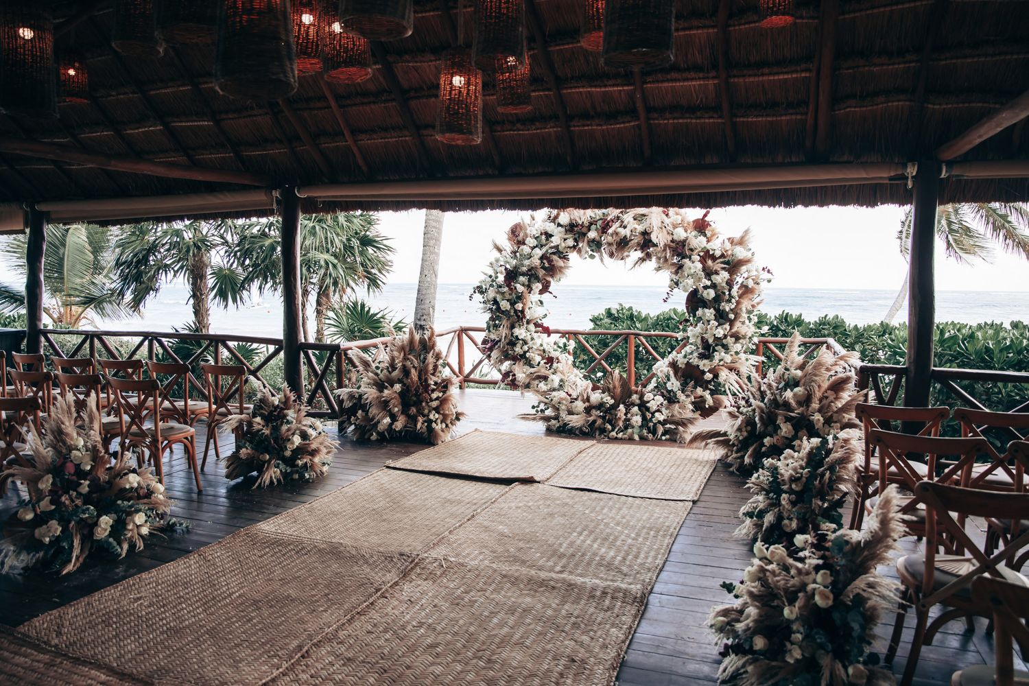 An elegant tropical wedding ceremony venue with a circular floral arch and pampas grass aisle decorations by the sea.