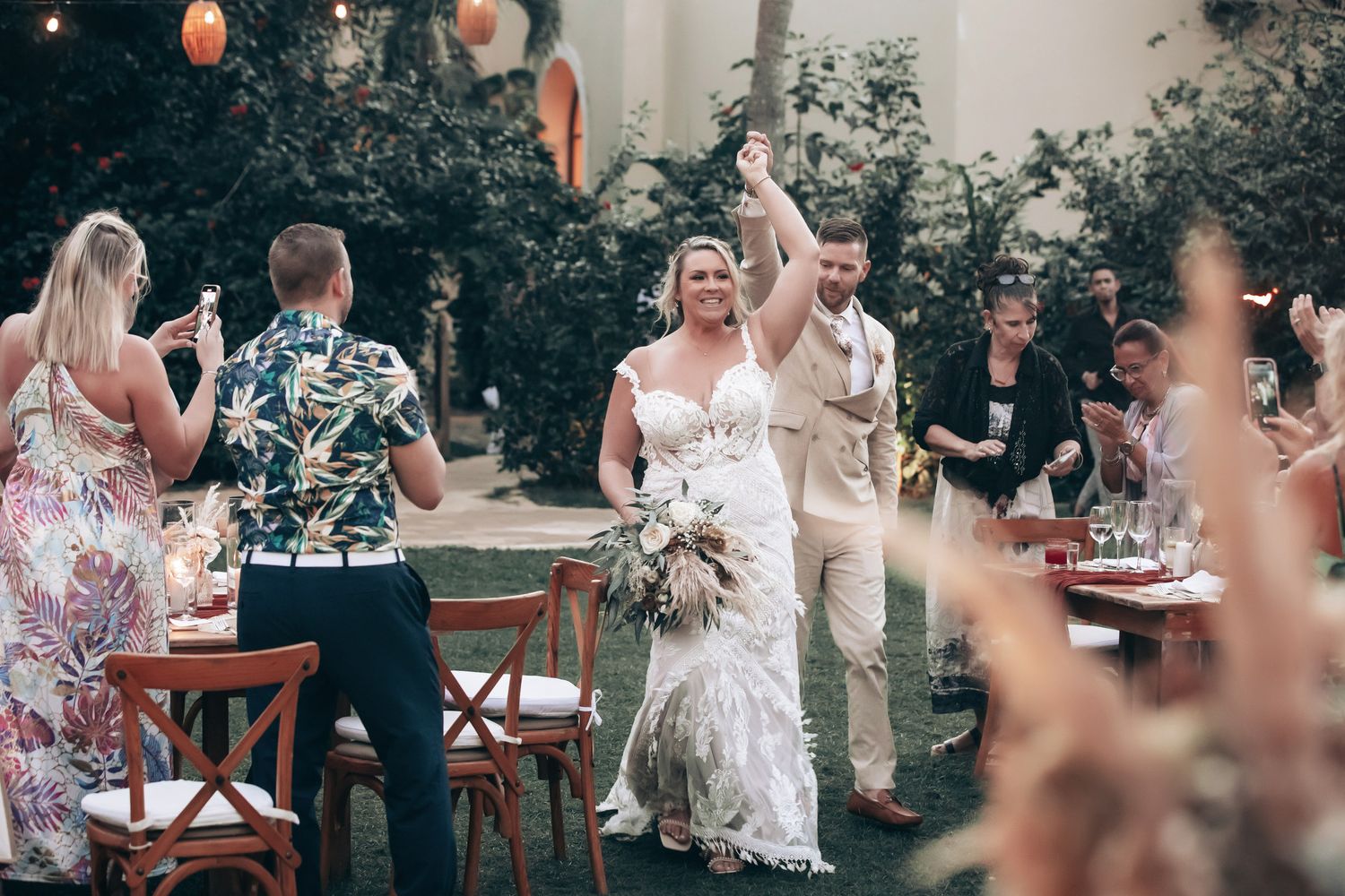 Bride and groom celebrate joyfully with raised hands at outdoor wedding reception with guests and decorated tables.