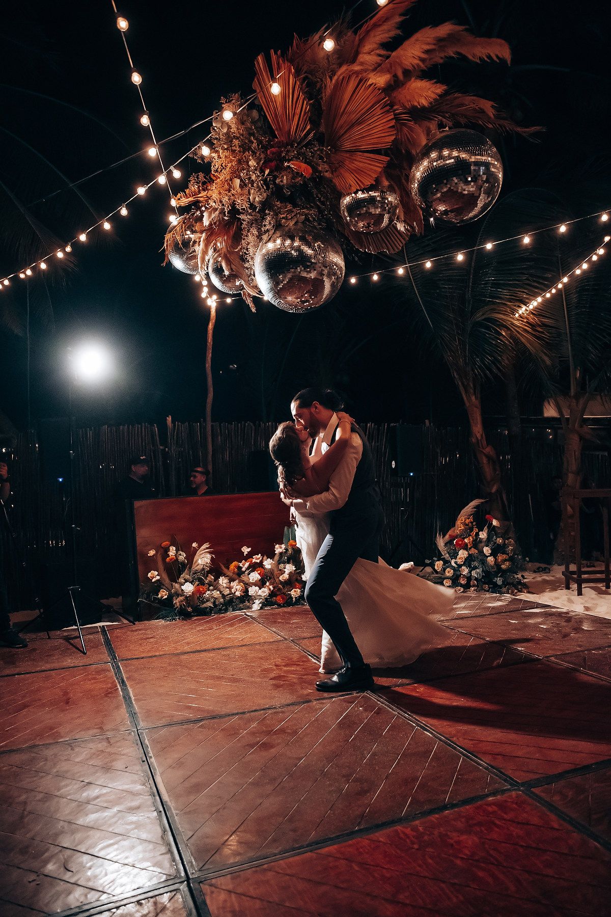 A couple performs their first dance on a wooden floor under romantic string lights and disco balls.