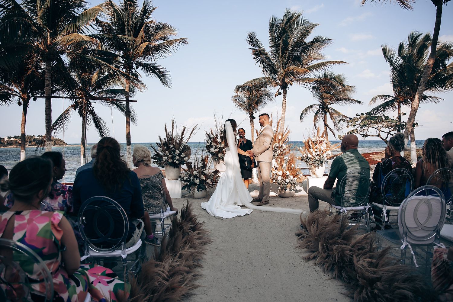 A beachside wedding ceremony with palm trees swaying in the breeze as guests gather on white chairs facing the ocean.