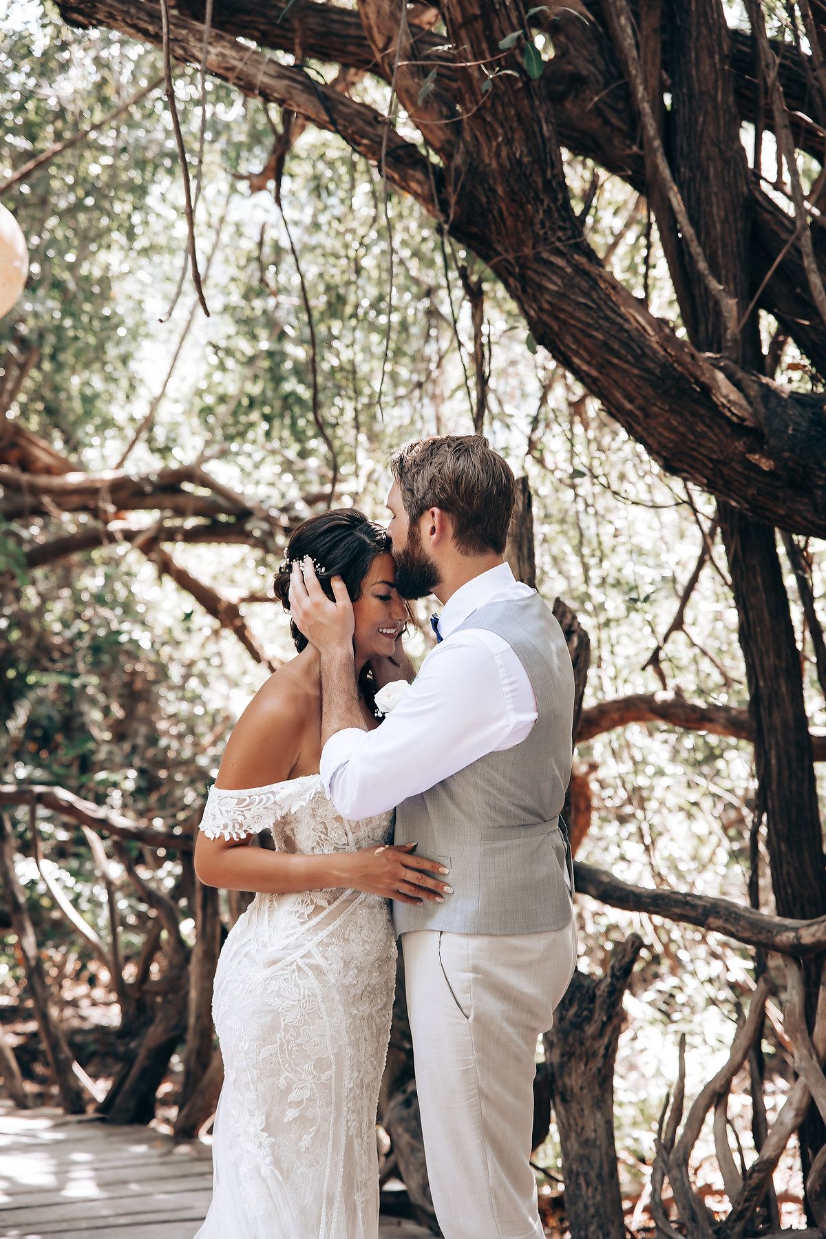 A couple embraces during their wedding photoshoot on a wooden walkway surrounded by trees.