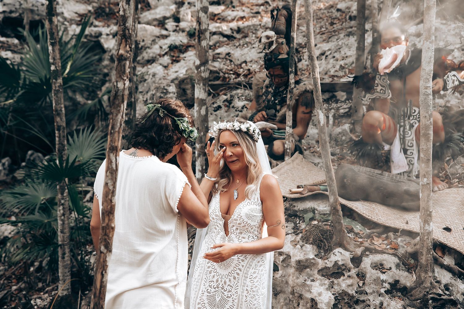 A couple in wedding attire embraces in a rustic outdoor setting with stone walls and greenery in the background.
