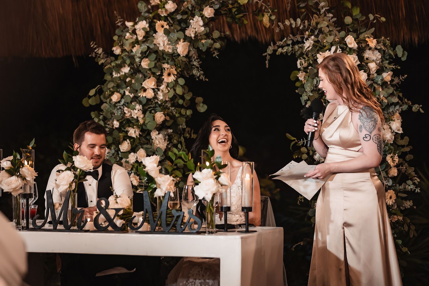 Bride and groom laughing at sweetheart table during wedding reception speech with floral arch backdrop.