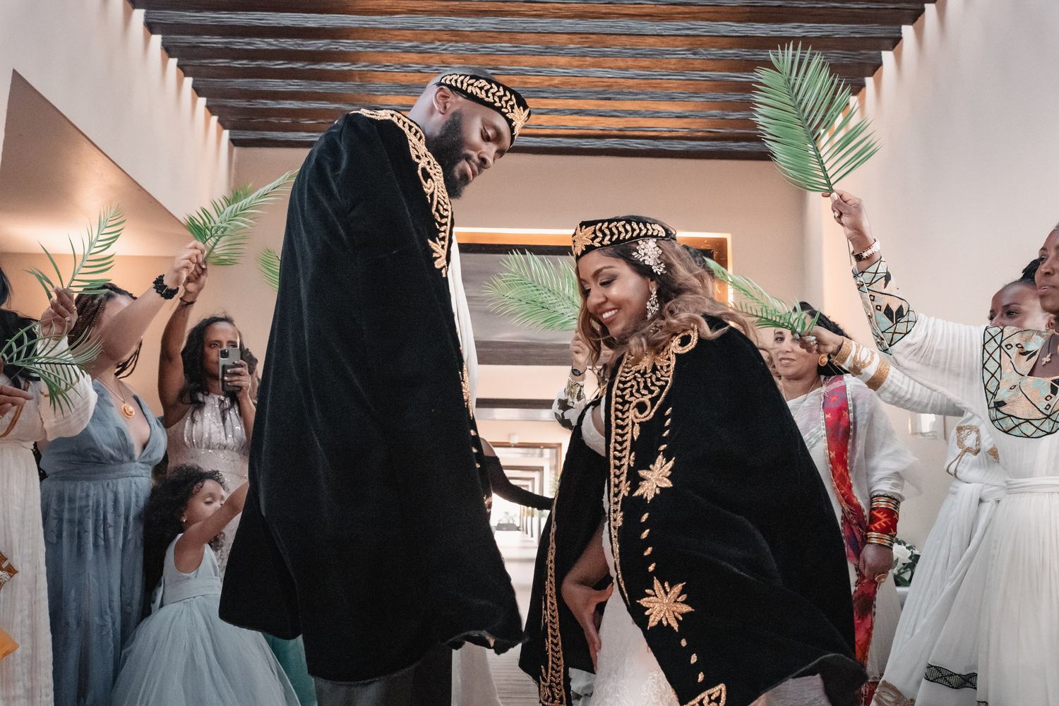 Couple dancing joyfully in ornate black and gold traditional Ethiopian wedding attire with palm-waving guests.