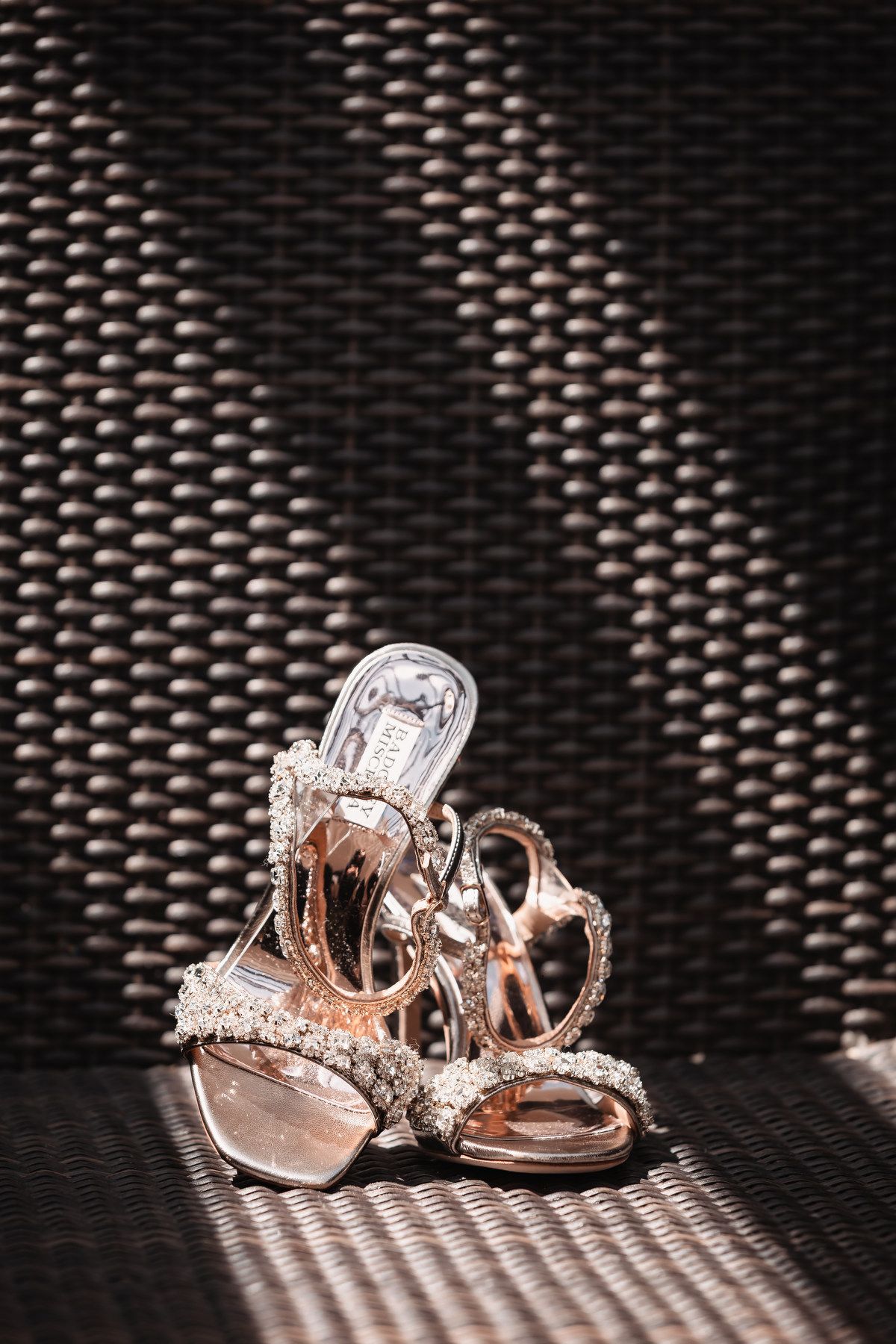 Elegant crystal-embellished rose gold strappy heels on woven rattan surface in natural light.