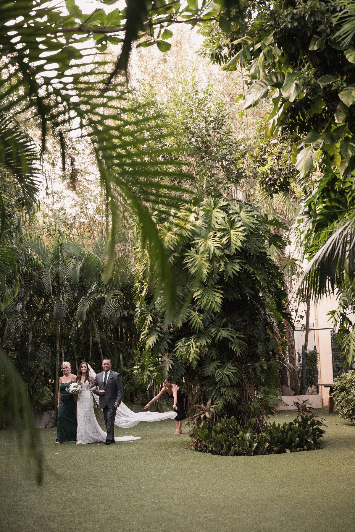 Bride walking down the aisle with her parents through a stunning tropical garden ceremony venue.
