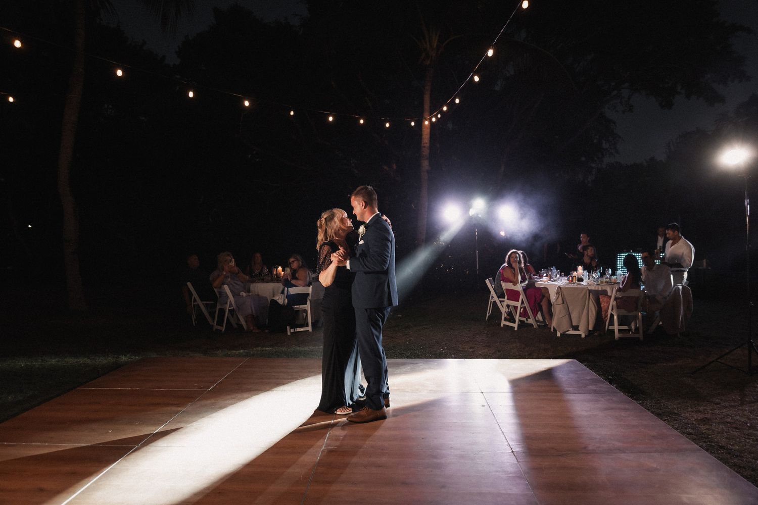 Groom dances with mother under string lights on outdoor dance floor at night reception.