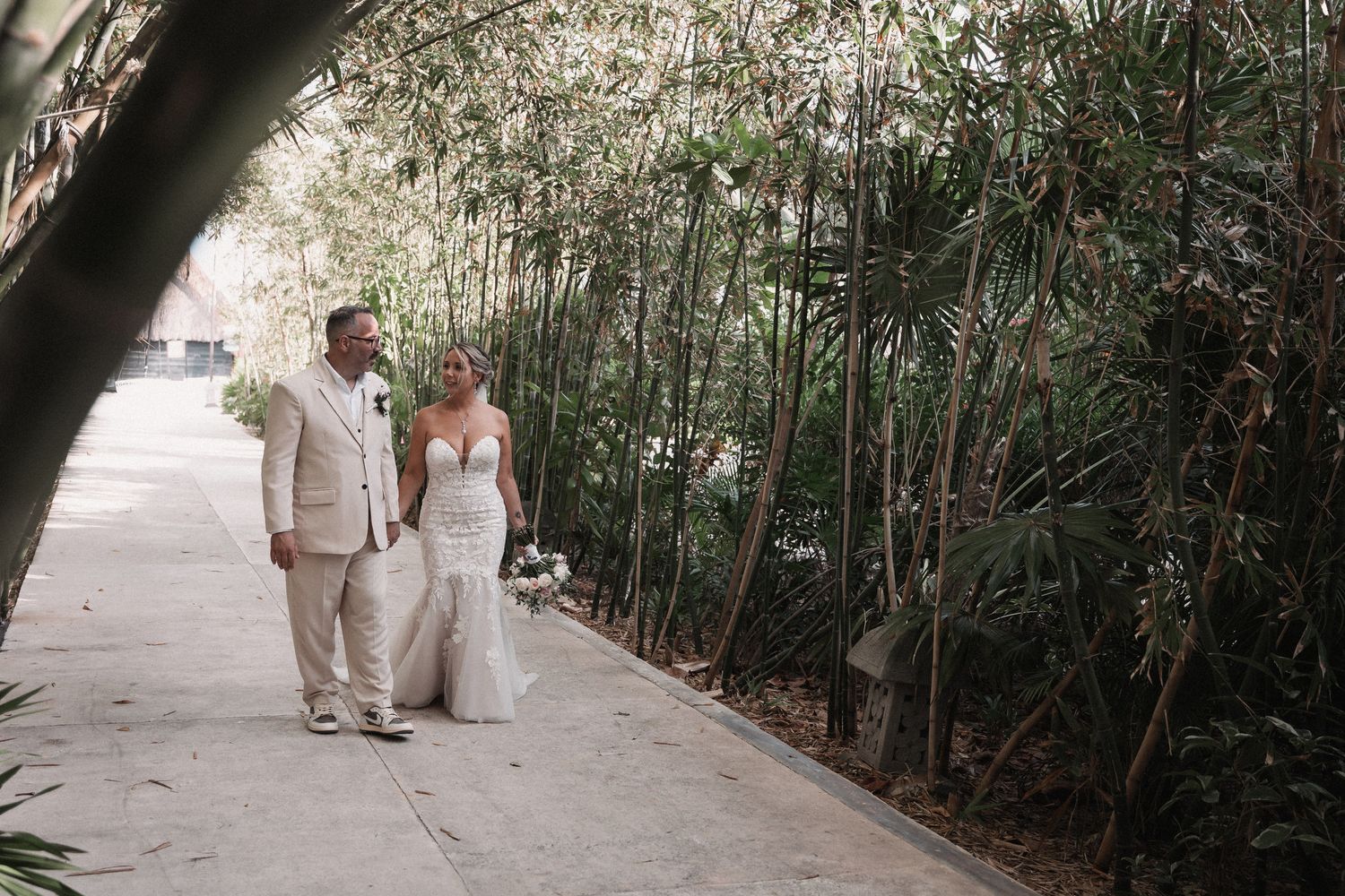 Bride and groom walking hand in hand along a lush bamboo-lined pathway on their wedding day.