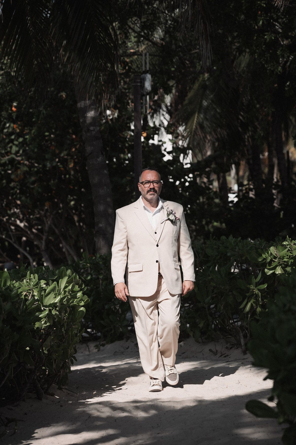 Groom in cream linen suit with boutonniere walking along sandy tropical palm tree lined pathway.
