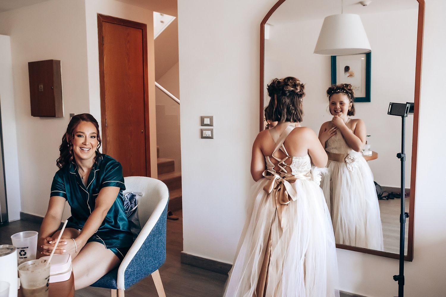 People getting ready in a bright room with a mirror, white dress, and chair visible.