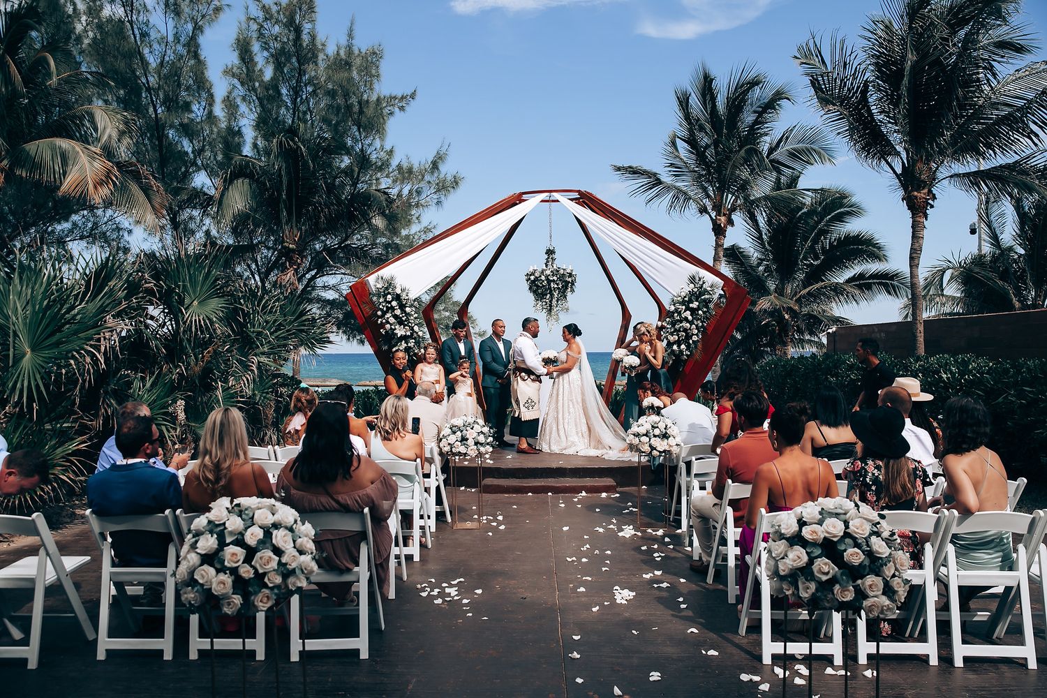 Beachside wedding ceremony setup with white chairs and tropical backdrop.