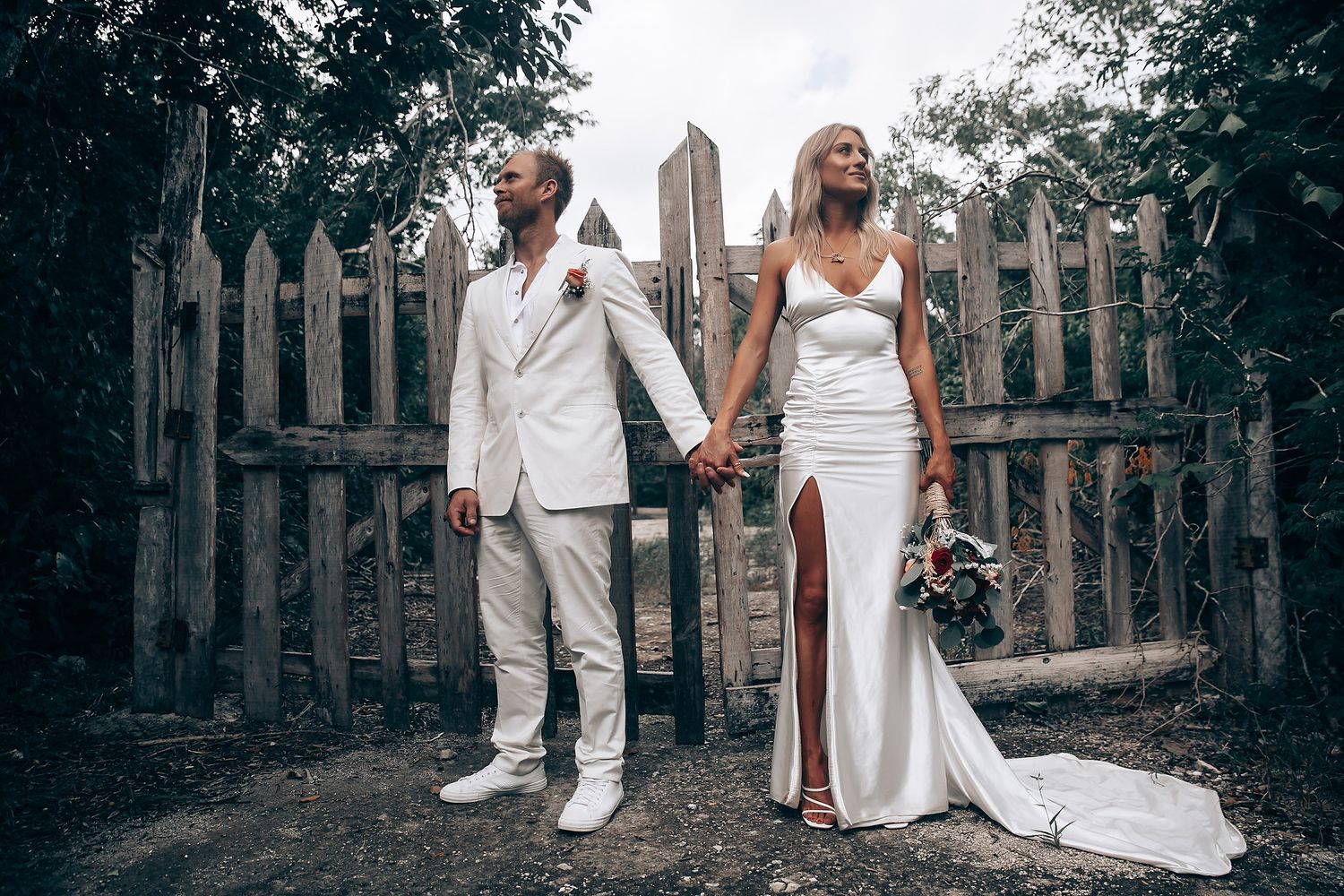 Wedding couple walking hand in hand along rustic wooden fence in garden setting.