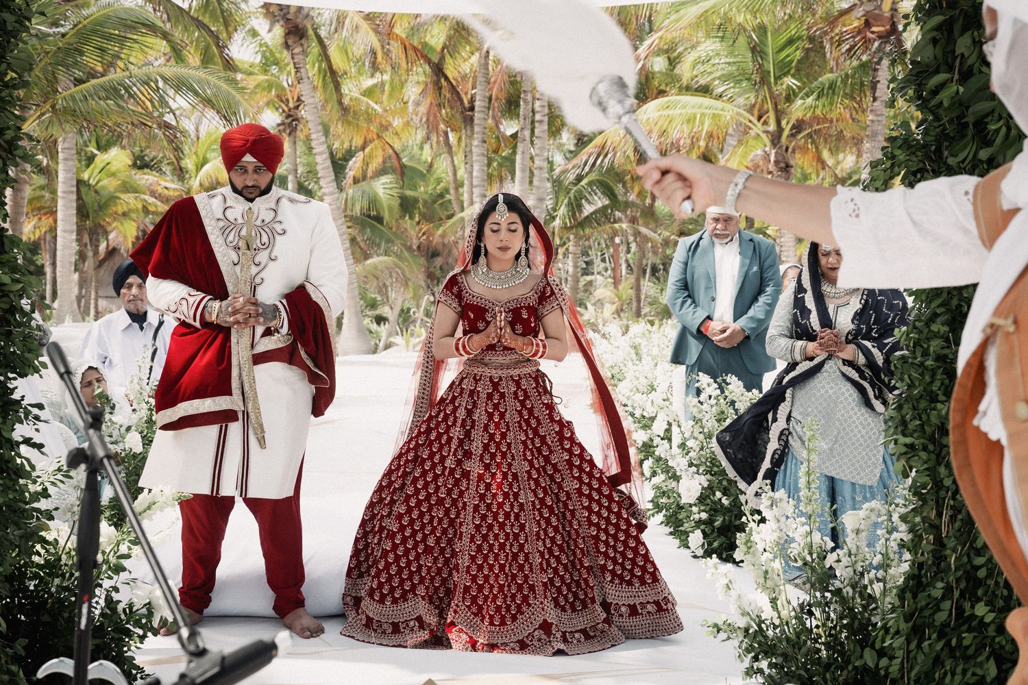 A Sikh bride in red lehenga walks with groom in white sherwani at a tropical outdoor wedding ceremony.