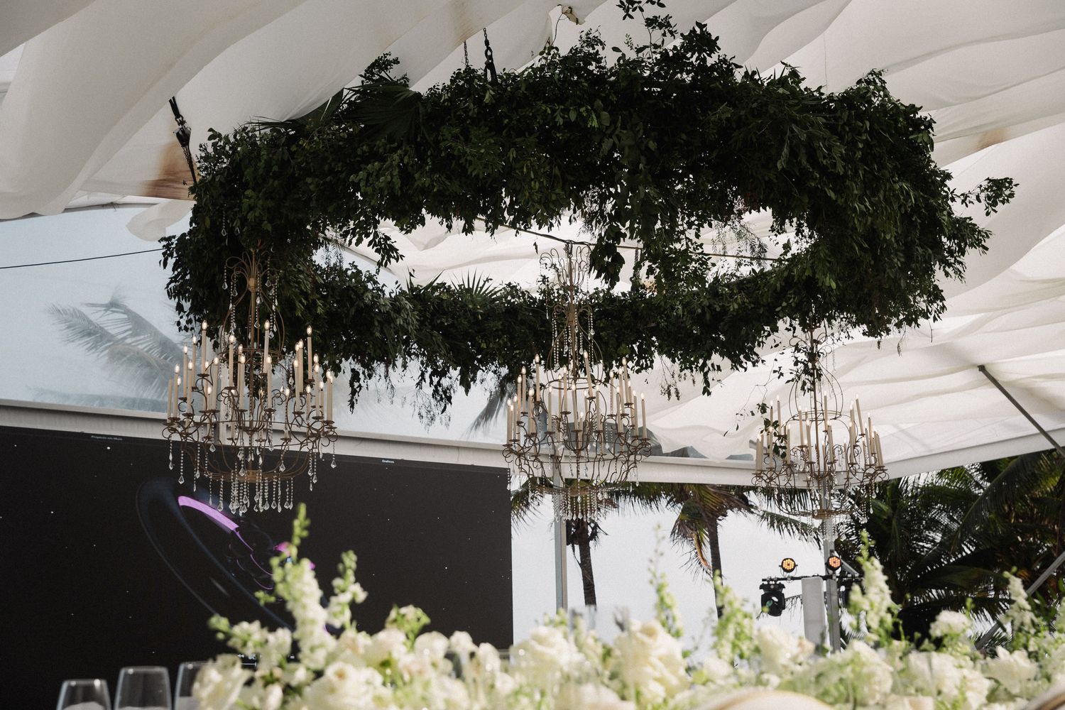 Circular green floral chandelier installation with crystal candelabras at elegant indoor wedding reception.