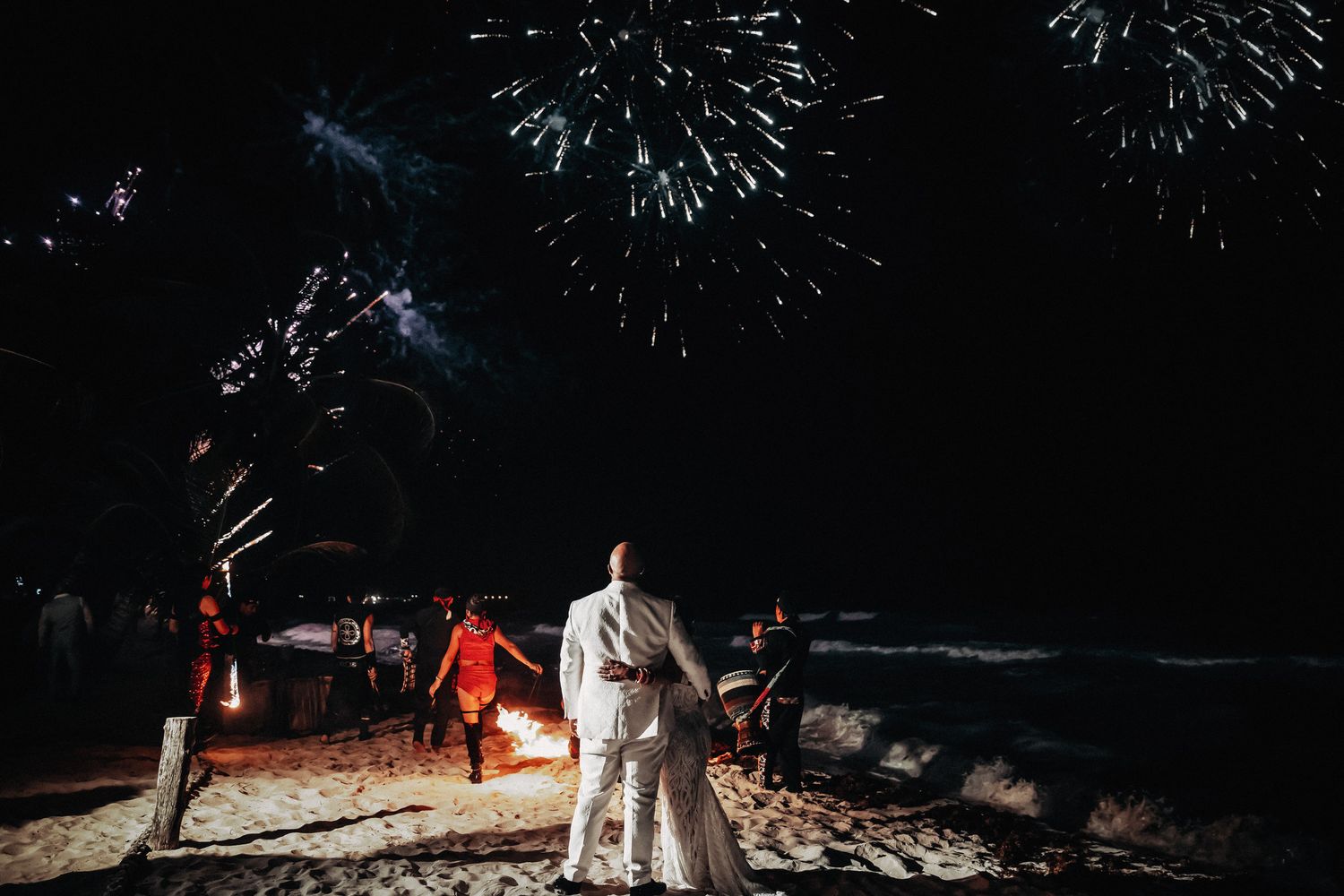 Bride and groom watch stunning fireworks display over ocean beach at romantic nighttime wedding celebration.