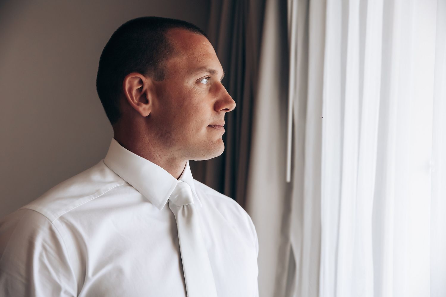 Groom in white dress shirt looks pensively out window during wedding preparations.