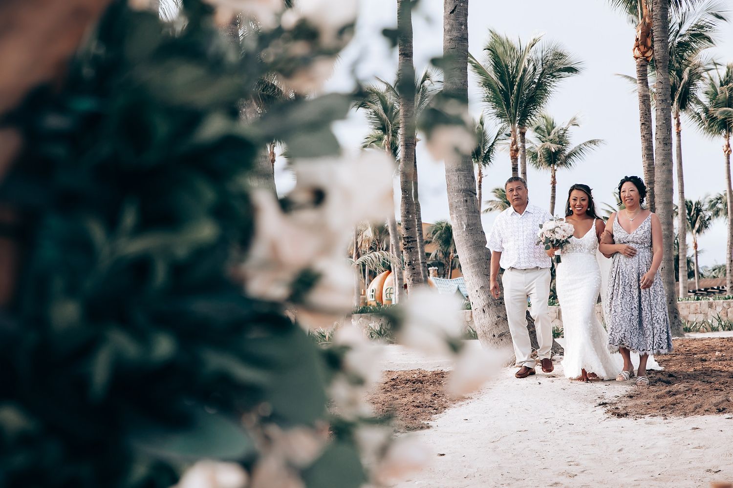Wedding party members walk down a palm-lined beach path in light colored formal attire.