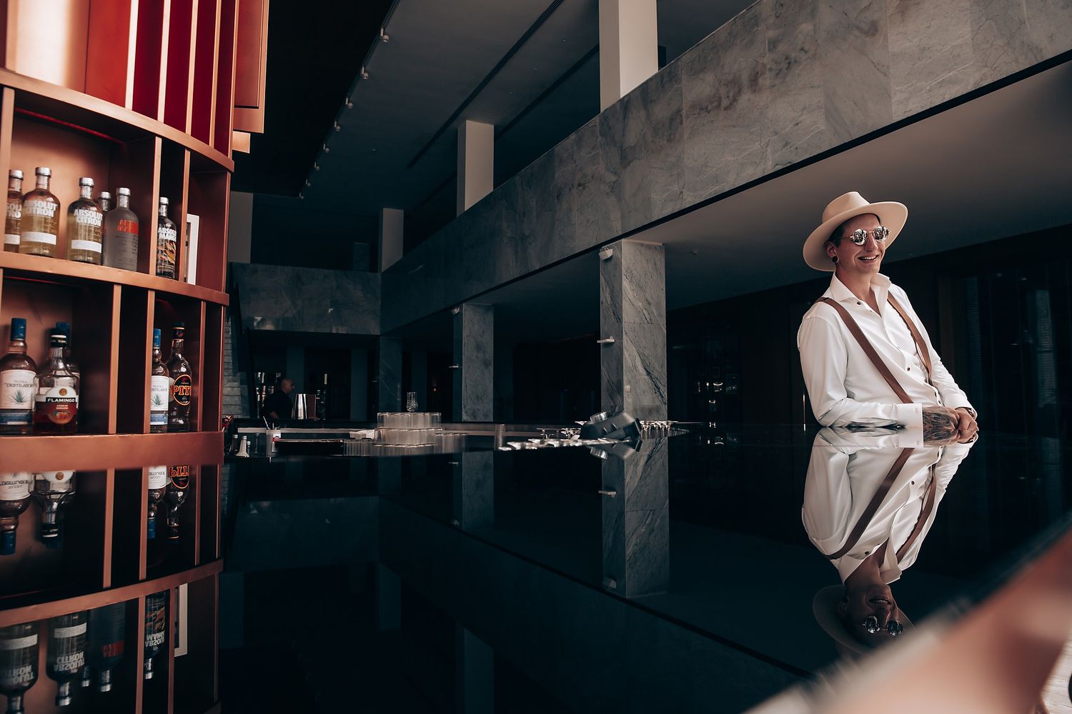 A moody interior shot featuring a white-suited figure reflected in a dark bar setting with bottles on display.
