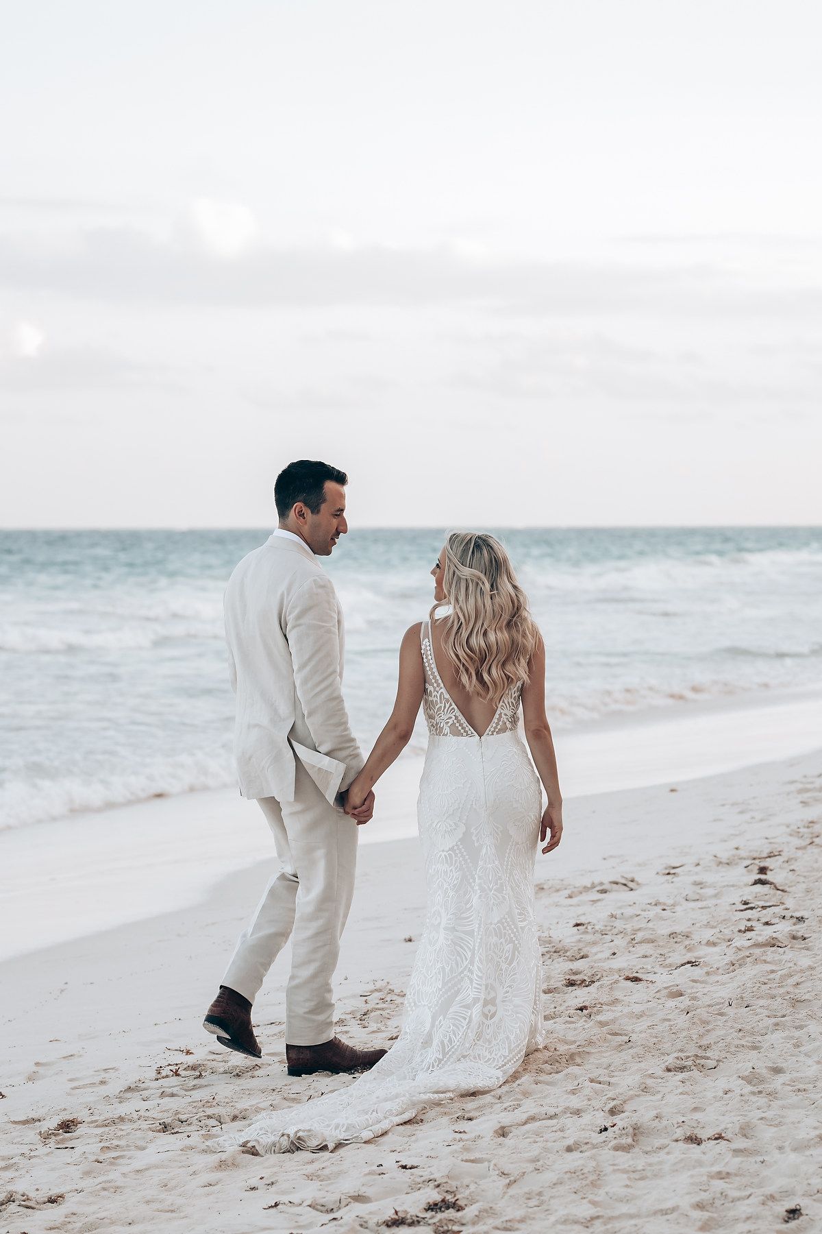 Couple in white attire walking along a serene beach shoreline.