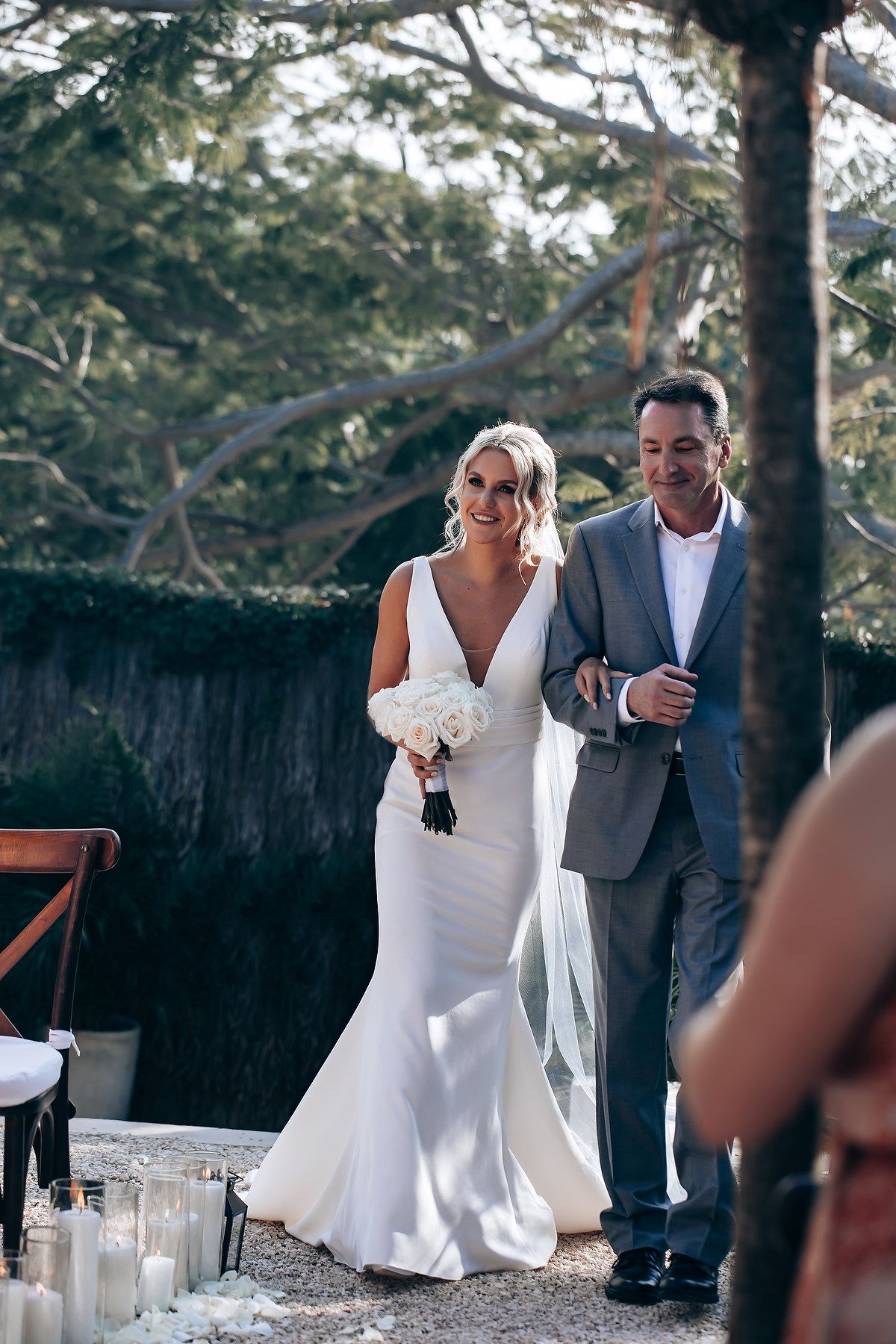 Outdoor wedding ceremony taking place under tall trees with white chairs and decorations.