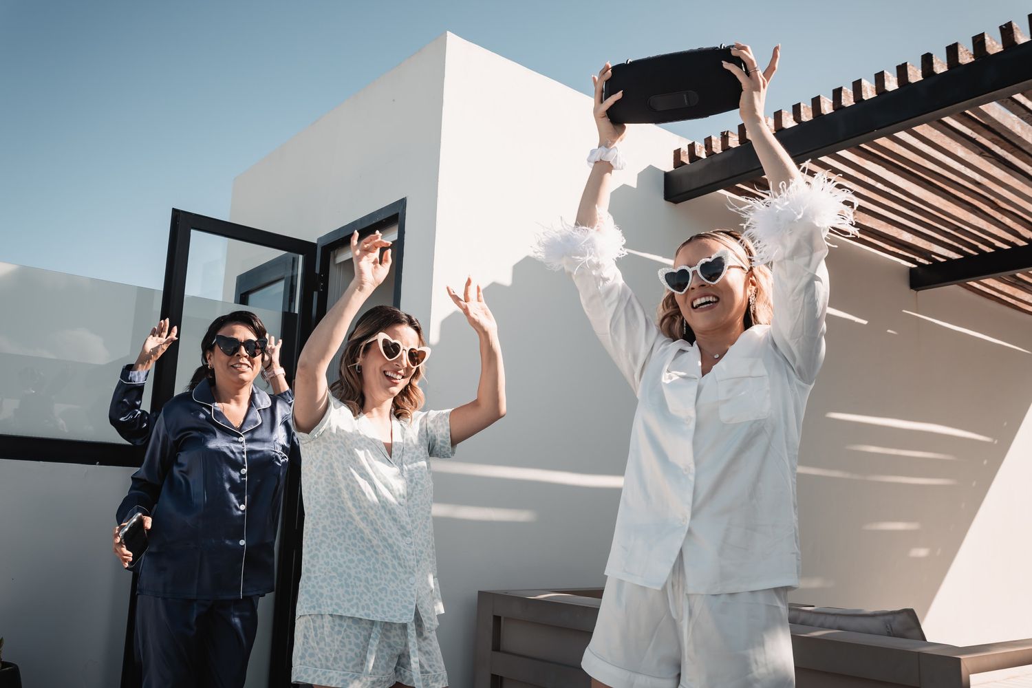 Three women in pajamas and sunglasses dancing outdoors, one holding a boombox overhead on a sunny day.