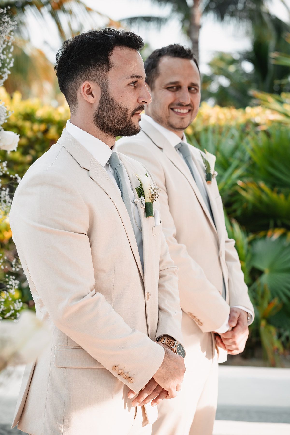 Two grooms in matching beige suits with boutonnieres stand at outdoor wedding altar surrounded by tropical greenery.