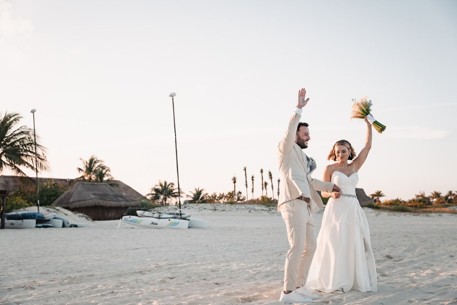Joyful newlyweds celebrate on tropical beach at sunset raising arms with bouquet.