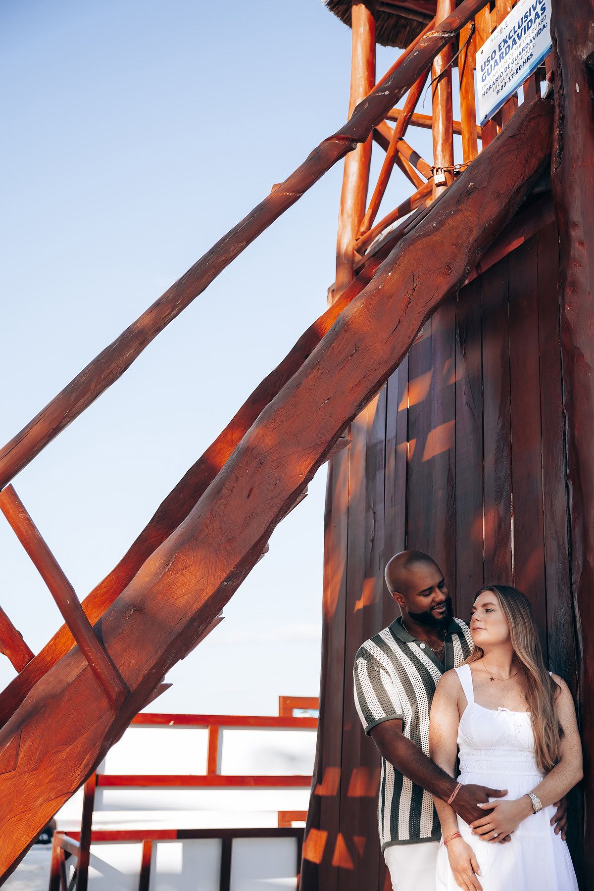 Interracial couple embracing near rustic red wooden structure outdoors under clear blue sky.